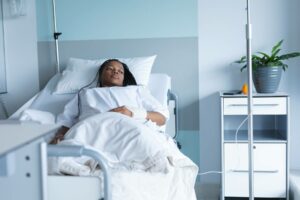 african american woman laying in hospital bed in white room, looking out towards window