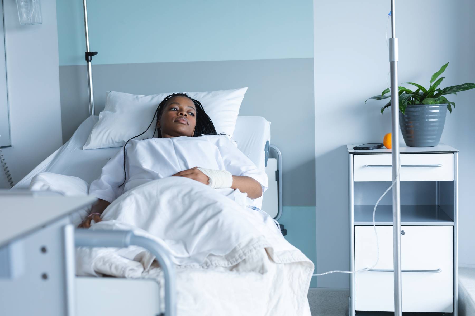 african american woman laying in hospital bed in white room, looking out towards window